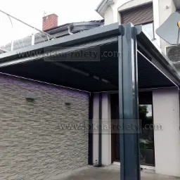 Nowoczesna, grafitowa konstrukcja z zadaszeniem lamelowym i oświetleniem LED, przylegająca do ściany budynku z dekoracyjną okładziną imitującą kamień, widoczna klimatyzacja i antena satelitarna...