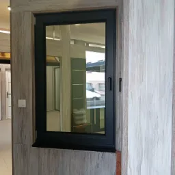 Ciemna rama okienna na tle drewnopodobnej ściany, widok na budynek i samochód przez szybę.