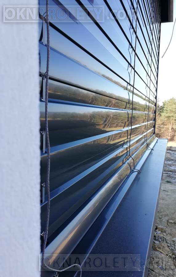 Zamknięte, poziome, metalowe żaluzje fasadowe w kolorze ciemnego grafitu, z widocznym sznurkiem sterującym i odbijającym się w powierzchni otoczeniem.