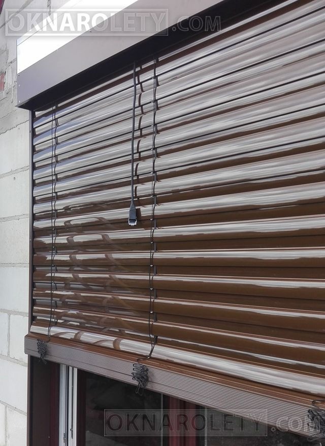 Zewnętrzna roleta w kolorze brązowym, częściowo opuszczona, z widocznym mechanizmem zwijania i czarnym sznurkiem do regulacji, zamontowana na oknie w budynku z białej cegły.