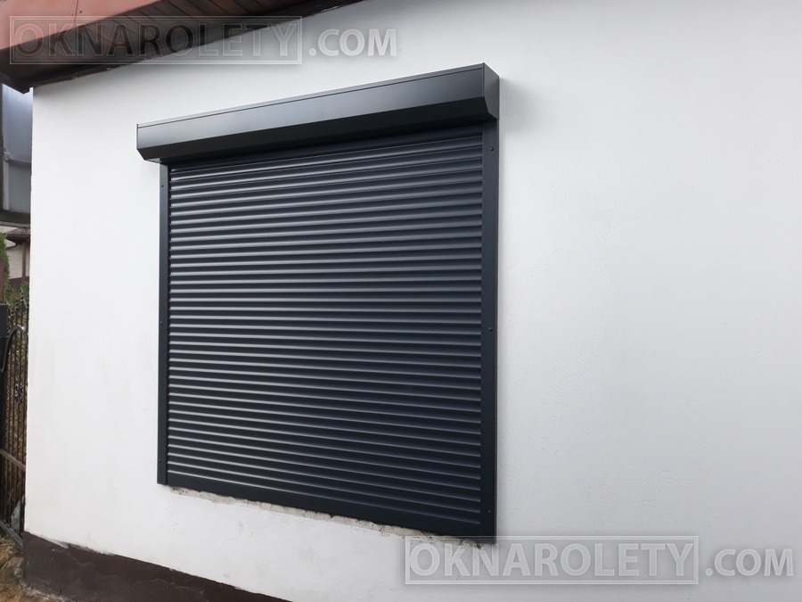 Zamknięta, grafitowa roleta zewnętrzna na oknie budynku z białą elewacją, widoczny fragment ogrodzenia po lewej stronie.