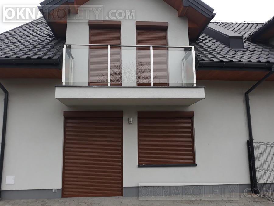 Dom z brązowymi roletami zewnętrznymi na oknach i drzwiach balkonowych, widoczne z zewnątrz, zamknięte, balkon z szklaną balustradą, szara elewacja, czarny dach.