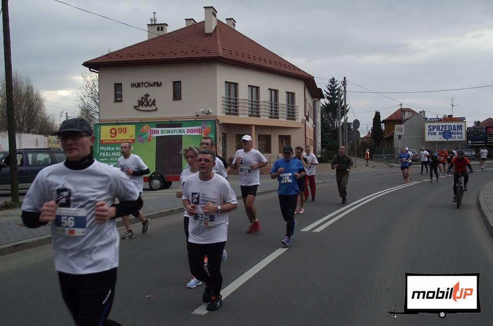 Bieg uliczny z uczestnikami w koszulkach z numerami, na tle budynku z reklamą 'Dni Bohatera Domu' na przyczepie.