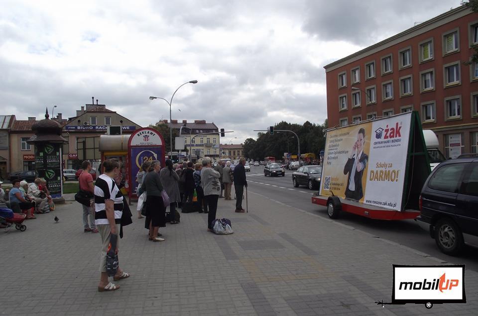 Przyczepa reklamowa z billboardem 'Szkoły za DARMO!' zaparkowana przy ruchliwej ulicy w mieście, obok przystanku autobusowego z oczekującymi pasażerami.