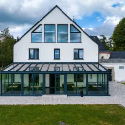 Nowoczesny biały dom z przeszklonym ogrodem zimowym, widoczne ciemne ramy okien i drzwi, kamienna ścieżka, trawnik, w tle las i częściowo budowa.