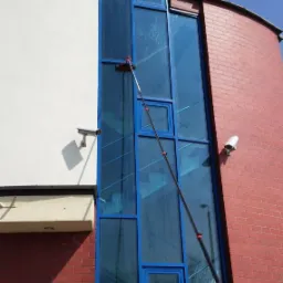 Czyszczenie okien na wysokości w budynku biurowym z użyciem teleskopowego kija i myjki, widoczna elewacja z czerwonej cegły i biały fragment fasady oraz okna z niebieskimi ramami.