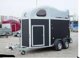 Szaro-czarna przyczepa laweta z pojedynczymi drzwiami i małym oknem, zaparkowana na zewnątrz; w tle widoczna czerwona przyczepa i budynek z reklamami.