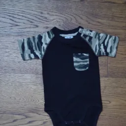 Czarne body dziecięce z krótkim rękawem w kamuflaż, leżące na drewnianej podłodze. Na ramionach zatrzaski, na piersi naszyta kieszonka w ten sam wzór.