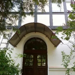 Wejście do budynku z zadaszeniem z poliwęglanu, widoczne przez gałęzie drzew. Po lewej stronie oparta o ścianę drabina. Biała elewacja z ciemnymi elementami konstrukcyjnymi.