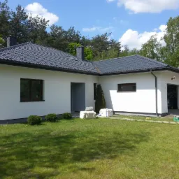 Biały dom z szarym dachem na tle zieleni. Widoczne okna z brązowymi ramami, trawnik i drzewa. Częściowo widoczne materiały budowlane przy wejściu.