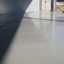 Jasnoszara, gładka posadzka w hali przemysłowej, z widocznym zarysowaniem na powierzchni i mocnym cieniem padającym z góry, w tle widoczne bale słomy pod zadaszeniem.