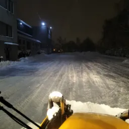 Żółta spycharka odśnieża oświetlony plac przed budynkiem w nocnej scenerii, widok z perspektywy operatora.