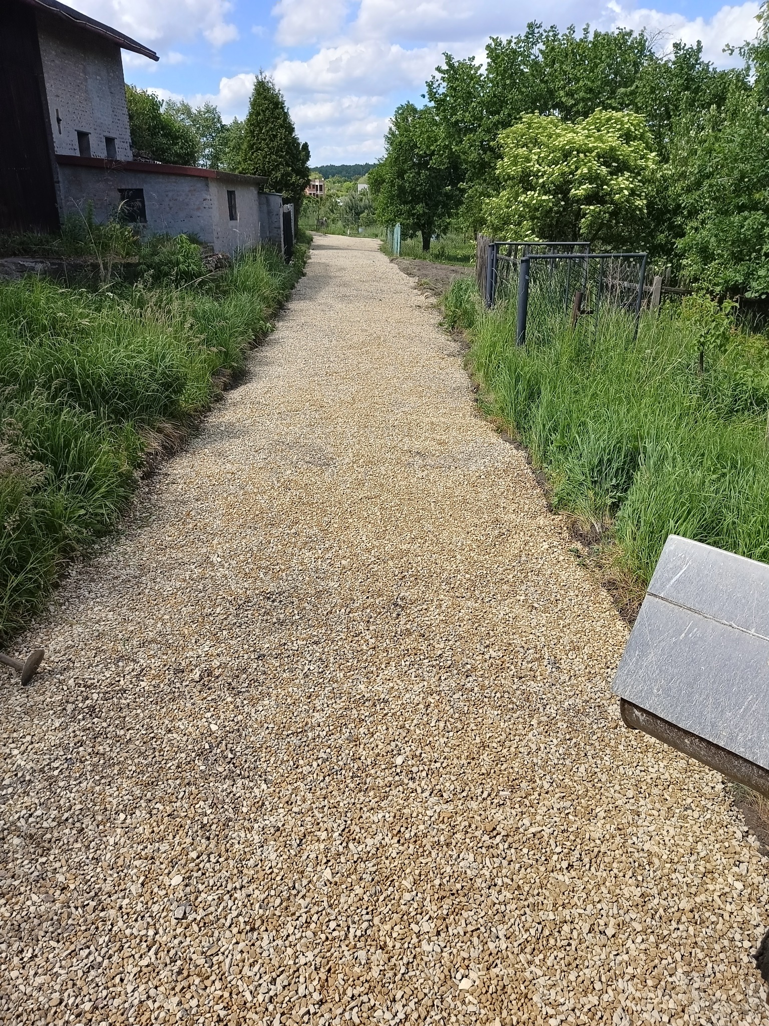 Długa, prosta droga dojazdowa wysypana drobnym, jasnym żwirem, otoczona bujną zielenią i ogrodzeniem, prowadząca w kierunku zabudowań i drzew w oddali.