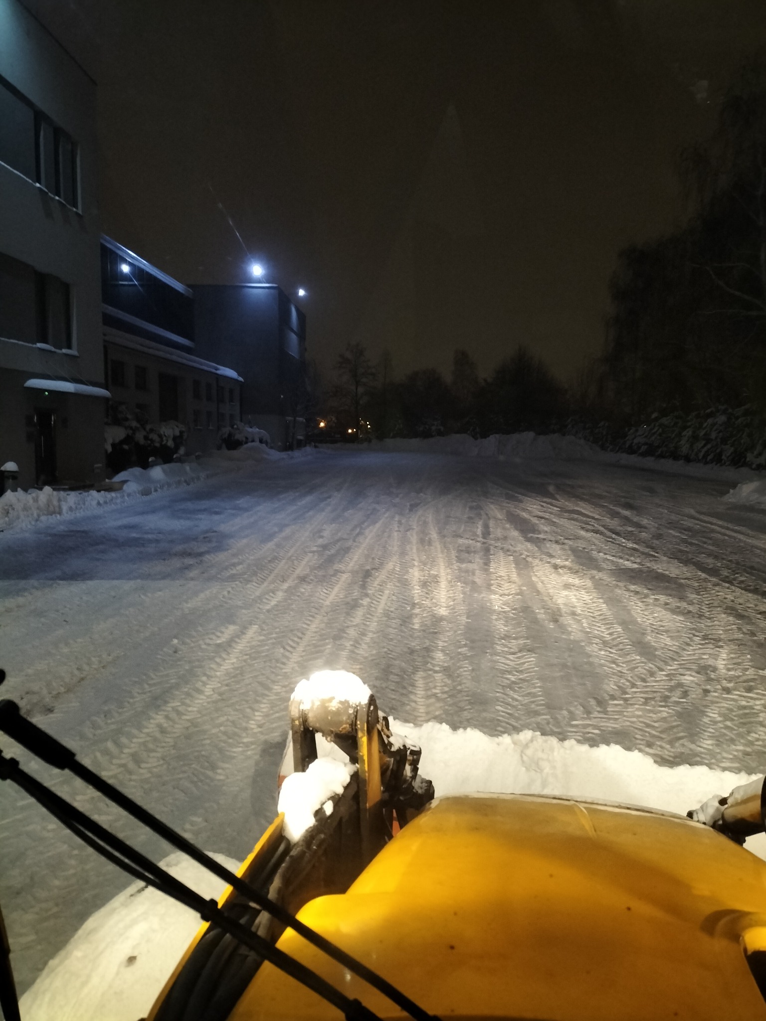 Żółta spycharka odśnieża oświetlony plac przed budynkiem w nocnej scenerii, widok z perspektywy operatora.