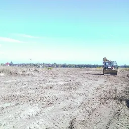 Teren po niwelacji z widocznym traktorem. Ślady opon na ziemi, w tle zabudowania i linia drzew. Błękitne niebo.