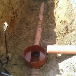 Wykop z brązowymi rurami kanalizacyjnymi i studzienką, widoczne narzędzia (łopata, szpadel) oraz ściany wykopu z ziemi. Brązowe rury kanalizacyjne.