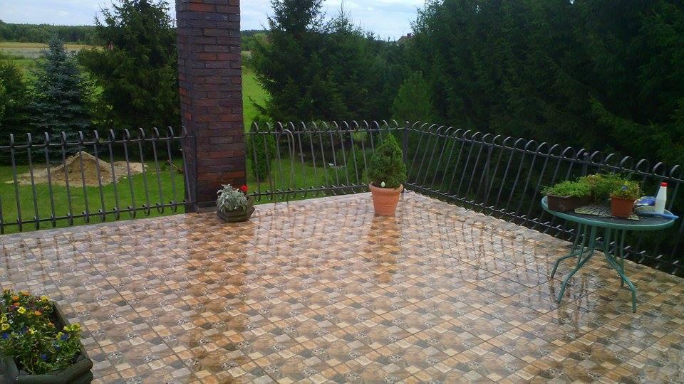 Balkon z mozaikową terakotą, czarna, ozdobna balustrada, widok na zieleń. Na stole rośliny w doniczkach i butelka. Słup z cegły klinkierowej.