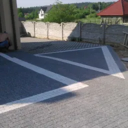 Ułożona kostka brukowa w szarym odcieniu z białymi pasami, tworzącymi wzór. Częściowo widoczny pracownik przy garażu i ogrodzenie posesji w tle.
