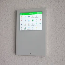 Panel sterowania systemem z zielonym interfejsem dotykowym zamontowany na ścianie o wyraźnej fakturze, wyświetlający ikony różnych funkcji, takich jak oświetlenie, żaluzje, wentylacja, muzyka...
