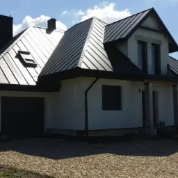 Nowoczesny dom jednorodzinny z ciemną, metalową dachówką, widoczne okno dachowe i komin, elewacja w trakcie wykończenia, podjazd wysypany żwirem, biały samochód dostawczy zaparkowany obok.