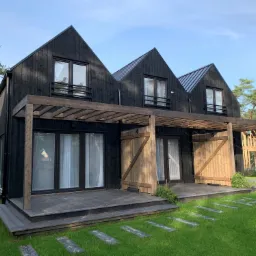 Nowoczesny dom z czarną drewnianą elewacją, trzema szczytowymi dachami i drewnianą pergolą nad tarasem, otoczony zielonym trawnikiem z kamiennymi płytami.