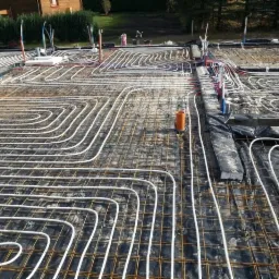 Rozłożone rury ogrzewania podłogowego na zbrojeniu fundamentu, widoczne pionowe rury instalacyjne i czarna folia izolacyjna.