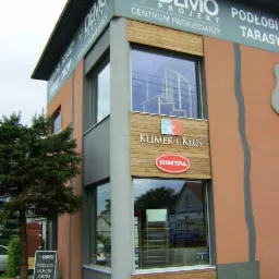 Fasada budynku z szyldami 'DEMO Projekt' i 'Klimek i Kłus', prezentująca ofertę podłóg, schodów i drzwi. W oknach ekspozycja produktów.