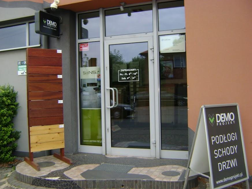 Wejście do salonu z ekspozycją podłóg i drzwi. Widoczny szyld firmy 'DEMO Projekt' oraz próbki różnych rodzajów podłóg drewnianych. Na drzwiach godziny otwarcia.