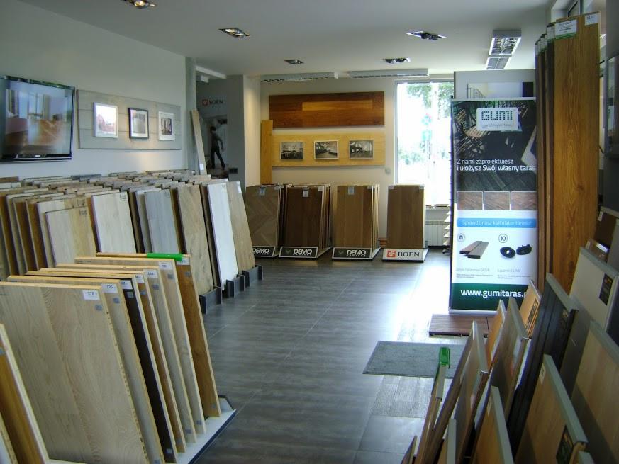 Salon z ekspozycją paneli podłogowych w różnych odcieniach drewna, z widocznym banerem reklamowym producenta GUMI i logotypami BOEN i DEMO. W tle widoczny fragment ekspozycji drzwi.