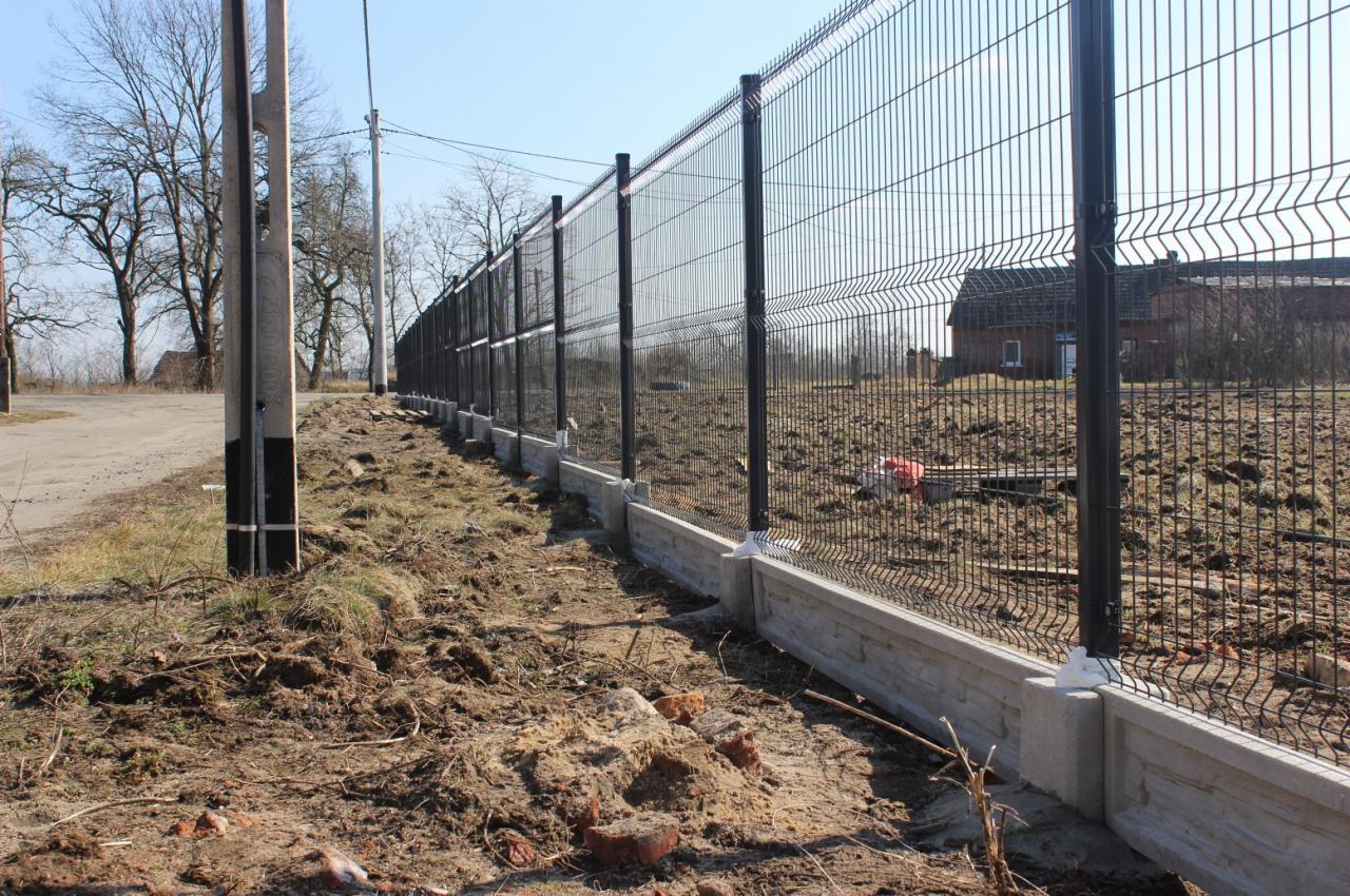 Długi, czarny płot panelowy na betonowej podmurówce w Gubinie. Widok z boku, na drugim planie budynek. Słup energetyczny po lewej stronie.