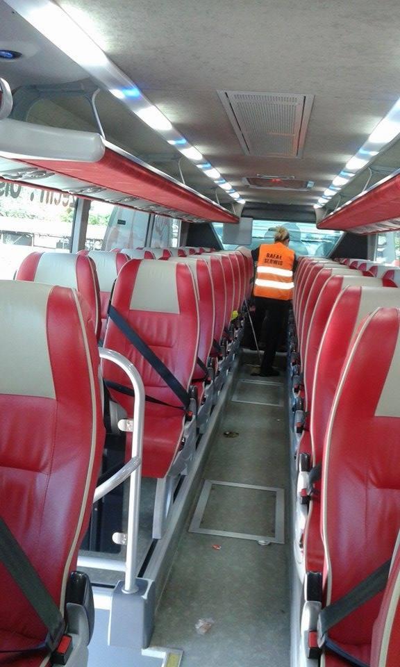 Wnętrze autobusu z czerwonymi fotelami; osoba w pomarańczowej kamizelce sprząta podłogę. Widoczny rząd siedzeń z pasami bezpieczeństwa i oświetlenie sufitowe.