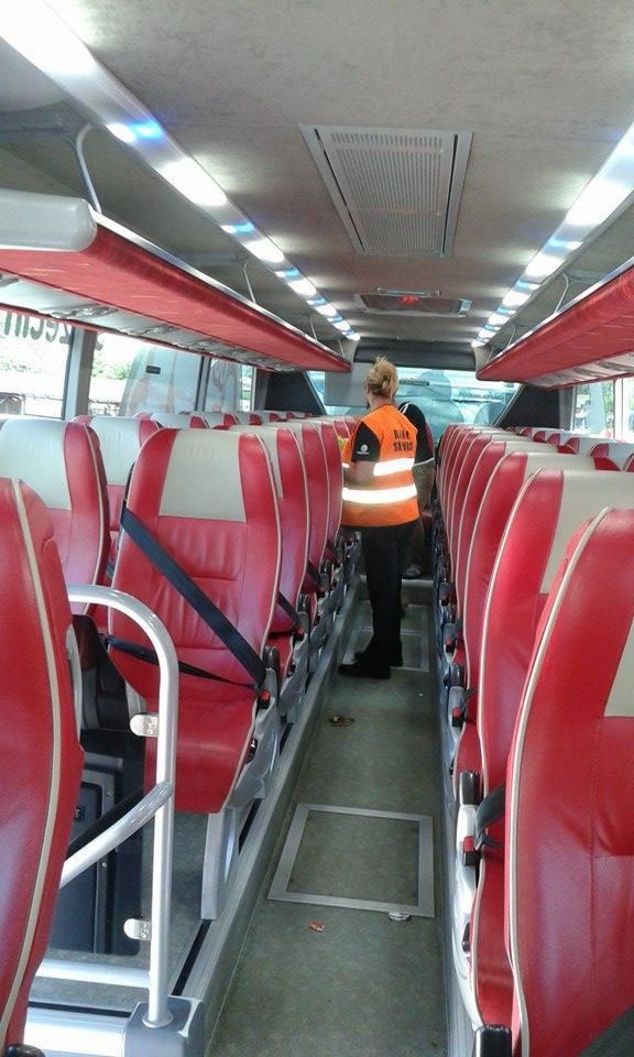 Wnętrze autobusu z rzędami czerwonych foteli. Osoba w pomarańczowej kamizelce stoi w przejściu. Widoczne oświetlenie sufitowe i okna.