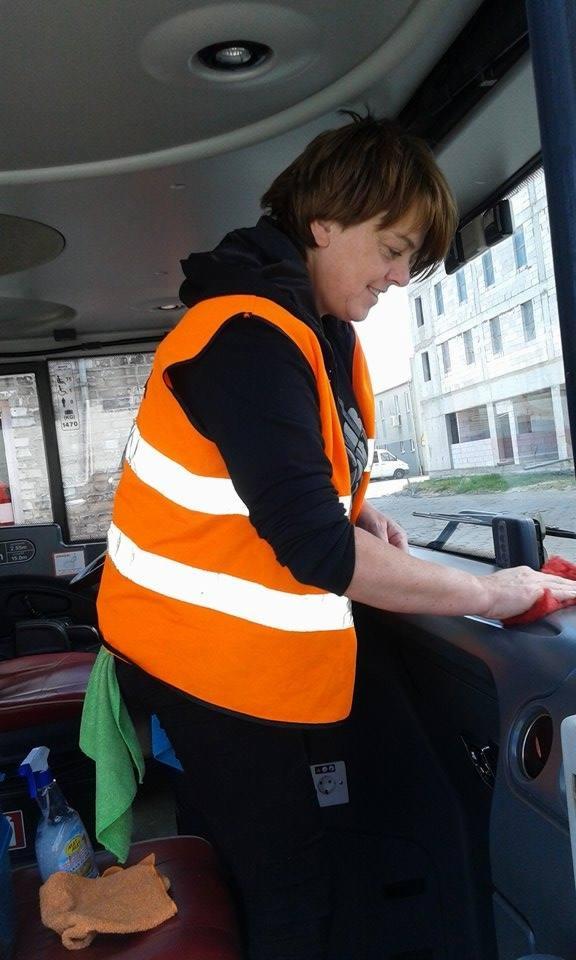 Pracownik w kamizelce odblaskowej czyści deskę rozdzielczą autobusu czerwoną ściereczką. Widoczny budynek w tle, butelka ze środkiem czyszczącym.
