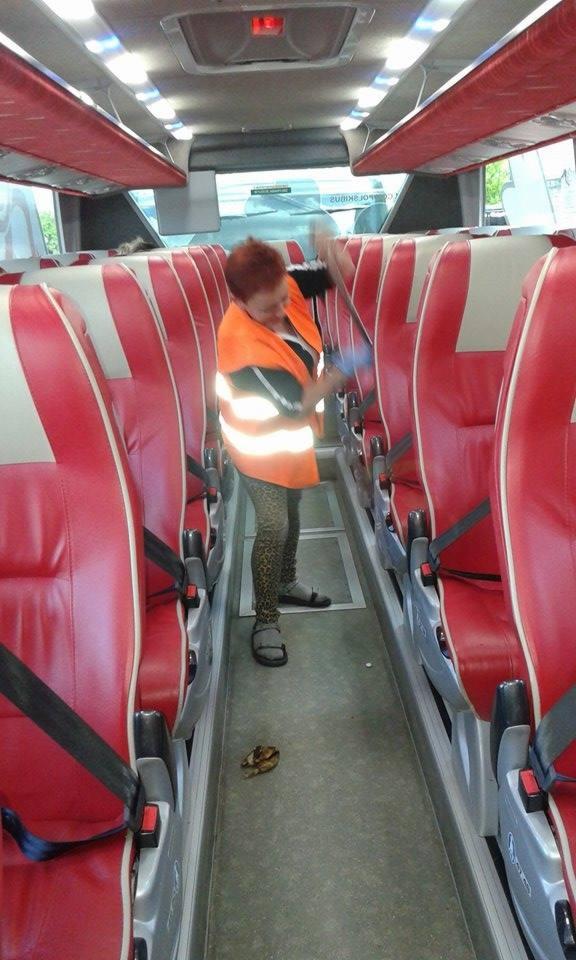 Kobieta w kamizelce odblaskowej czyści wnętrze autobusu mopem, widoczne czerwone fotele i fragment podłogi. W tle szyba autobusu.