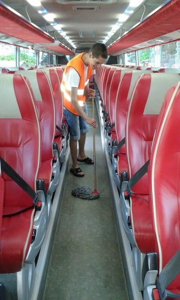 Mężczyzna w kamizelce odblaskowej myje podłogę w autobusie mopem sznurkowym. Czerwone fotele z pasami bezpieczeństwa po obu stronach przejścia.