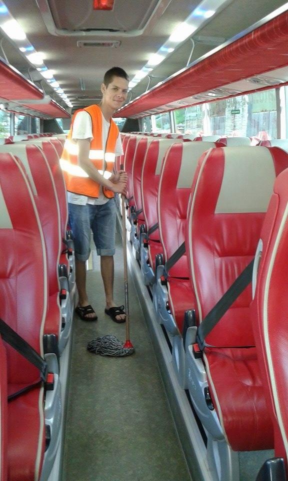 Mężczyzna w kamizelce odblaskowej myje podłogę w autobusie mopem. Wnętrze autobusu z czerwonymi siedzeniami. Widok z perspektywy środka pojazdu.