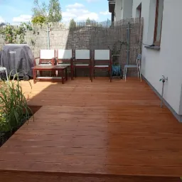 Nowoczesny taras z drewna kompozytowego w odcieniach brązu, z meblami ogrodowymi i elementami zieleni, przylegający do białego budynku z widocznym kranem na ścianie.