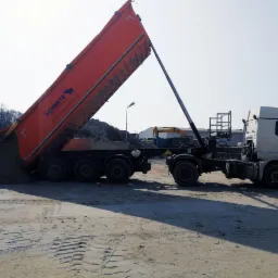 Pomarańczowa wywrotka marki Schmitz w trakcie wysypywania szarego kruszywa na placu budowy, w tle widoczna koparka.