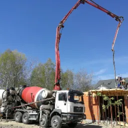 Betonowanie stropu domu jednorodzinnego z wykorzystaniem pompy do betonu MAN i betonomieszarki, widoczne szalunki i pracownicy budowlani na tle błękitnego nieba.