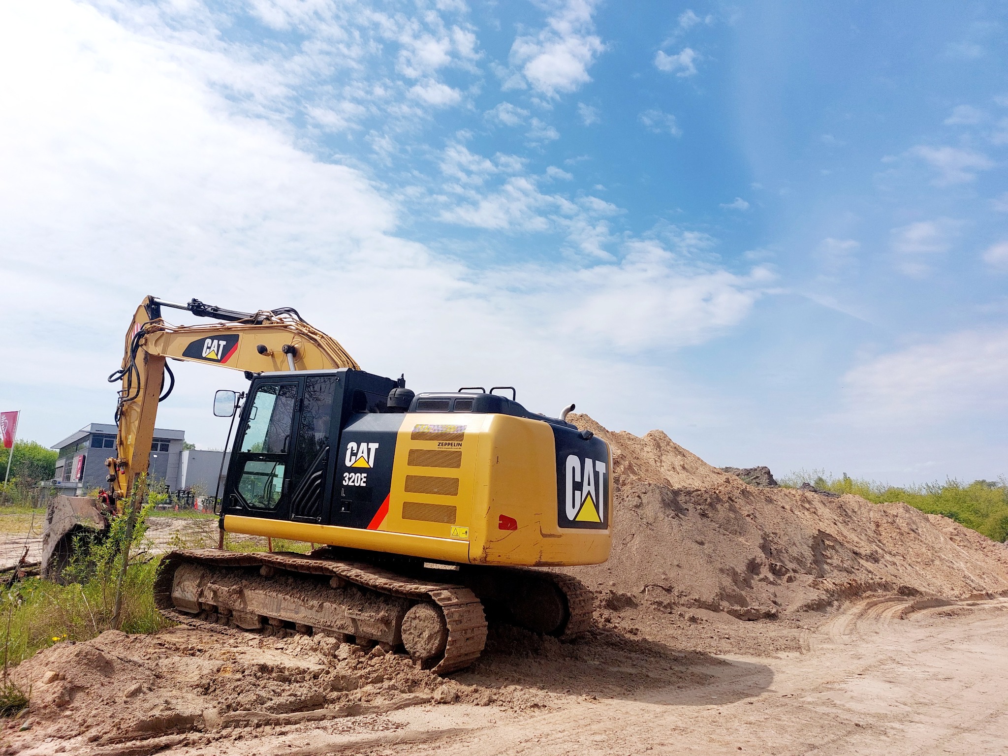 Żółto-czarna koparka CAT 320E stoi na piaszczystym terenie z hałdą ziemi w tle, pod błękitnym niebem z białymi chmurami.