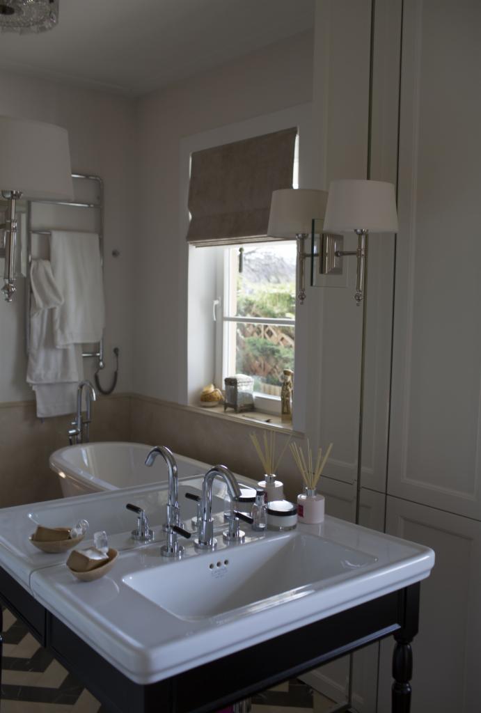 Elegancka łazienka z umywalką nablatową, bateriami w stylu retro i widokiem na zieleń za oknem. Blat w kolorze białym, szafka czarna. Lampa ścienna z abażurem.