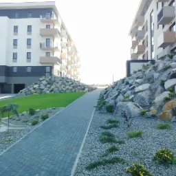 Widok na osiedle mieszkaniowe z brukowaną ścieżką, skalniakami i zielenią, jasne oświetlenie.