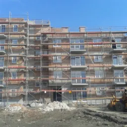 Budowa bloku mieszkalnego w trakcie wznoszenia, widoczne rusztowania, cegły i sprzęt budowlany na placu budowy, błękitne niebo w tle.