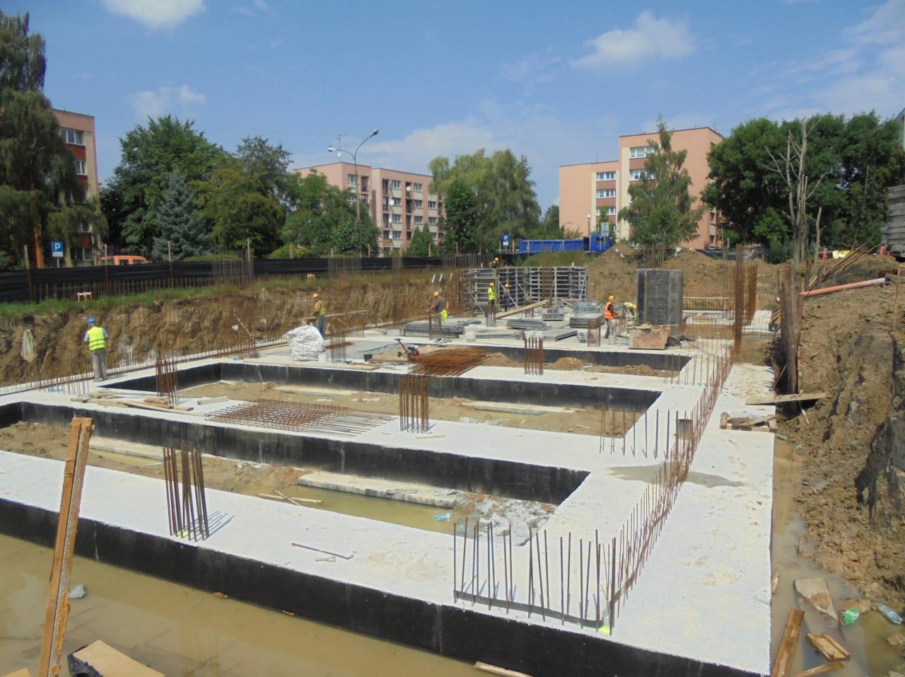 Widok z góry na wylane fundamenty pod budynek mieszkalny, z wystającymi prętami zbrojeniowymi, na tle bloków mieszkalnych i pracujących robotników w strojach ochronnych.