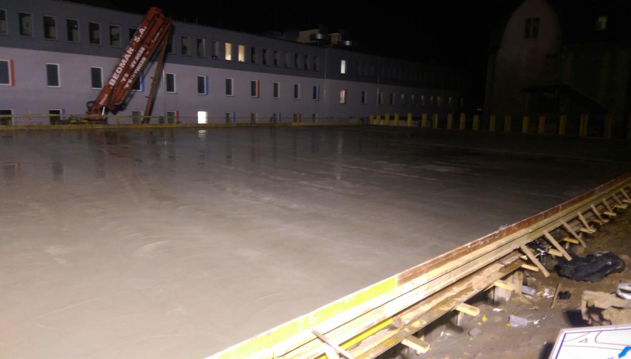 Wylany, świeży beton na dużej powierzchni, z widocznym podziałem na sekcje i podporami. W tle budynek z oknami i wysięgnik pompy do betonu.