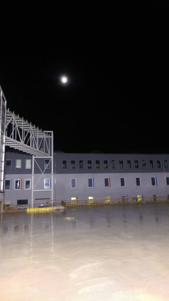 Nocny widok na plac budowy z konstrukcją stalową i budynkiem w tle, odbijający światło księżyca w mokrej nawierzchni.