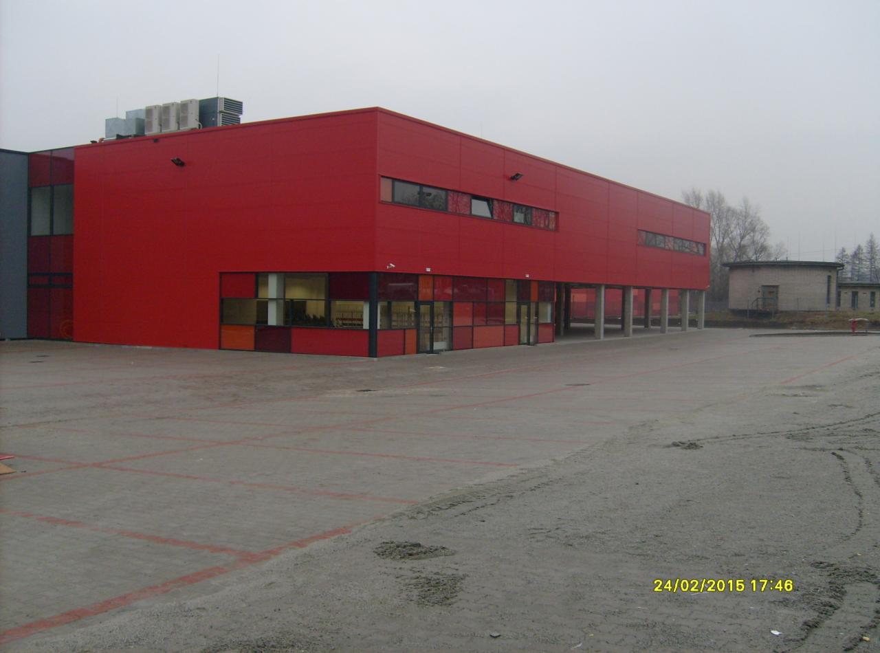 Czerwony budynek z betonowym placem. Widoczne linie wyznaczające miejsca parkingowe i mały budynek w tle. Pochmurna pogoda.