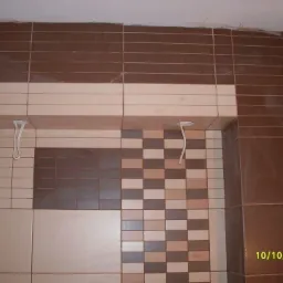 Wykończona ściana z brązowo-beżowych płytek ceramicznych, ułożonych w różne wzory. Widoczne przewody elektryczne wystające ze ściany. Data w prawym dolnym rogu.