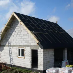 Niedokończony dom z białego pustaka, z częściowo wykonanym dachem. Drabina oparta o ścianę, worki z materiałami budowlanymi na ziemi. Widok na budowę w słoneczny dzień.