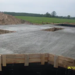 Zalany betonem fundament z drewnianym szalunkiem na czarnej izolacji. W tle pole i kopiec ziemi. Ujęcie z góry, lekko z prawej strony. Data 25/12/2008 w prawym dolnym rogu.